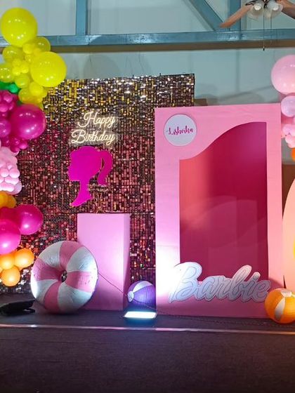 A vibrant, tropical Barbie-themed birthday party. This setup features a pink "doll box" photo booth, a sequin backdrop, and colorful balloon clusters with palm tree cutouts.