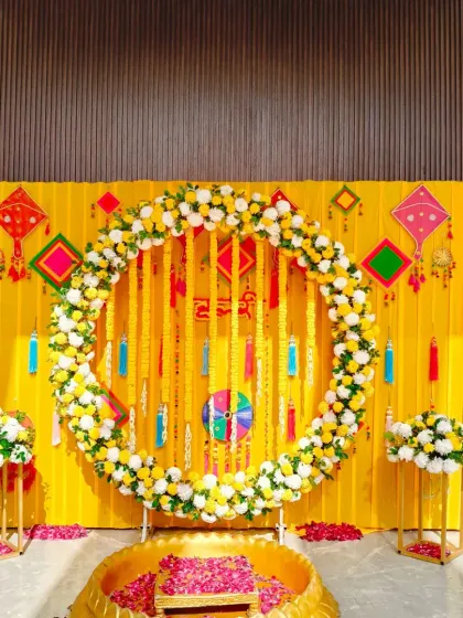 A beautiful Haldi backdrop featuring a large floral ring of marigolds and white flowers against a bright yellow curtain. The setup is complete with a traditional brass urli for the ceremony.