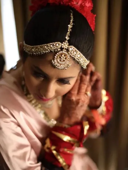 A candid moment of a bride getting ready, putting on her earrings. Her hair is styled in a neat bun, adorned with a beautiful matha patti, ready for her ceremony.