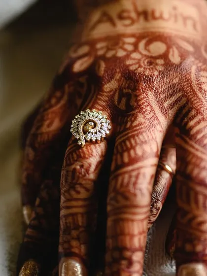 A close up of the bride's hand, showing off her engagement ring and the rich color of her mehendi, with the groom's name written on it.