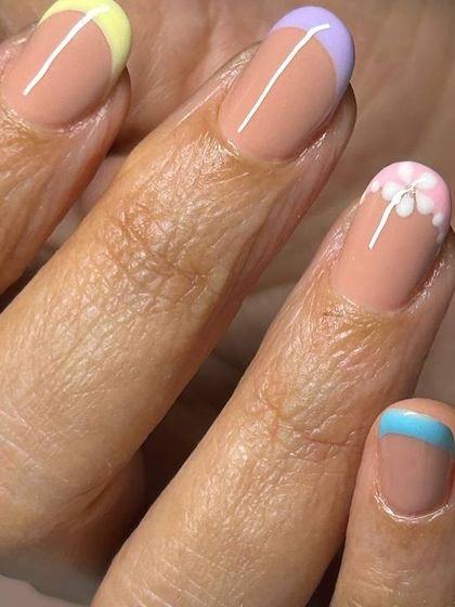 A playful and colorful take on the French manicure. Each nail features a different pastel-colored tip, with the ring finger adorned with a simple, delicate flower.