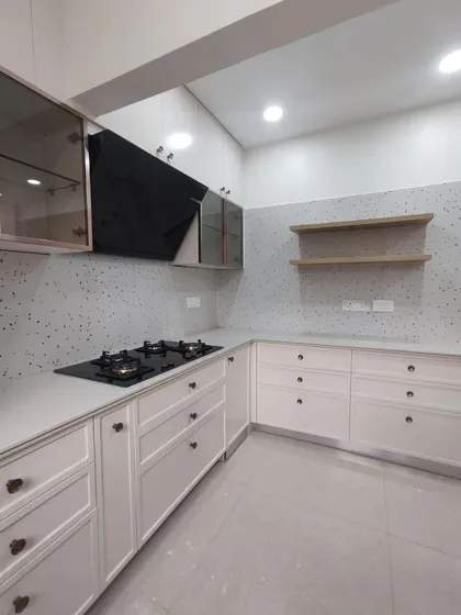 A clean and minimalist kitchen with white cabinets, a terrazzo-style backsplash, and open wooden shelves. The black hob and chimney provide a striking contrast.