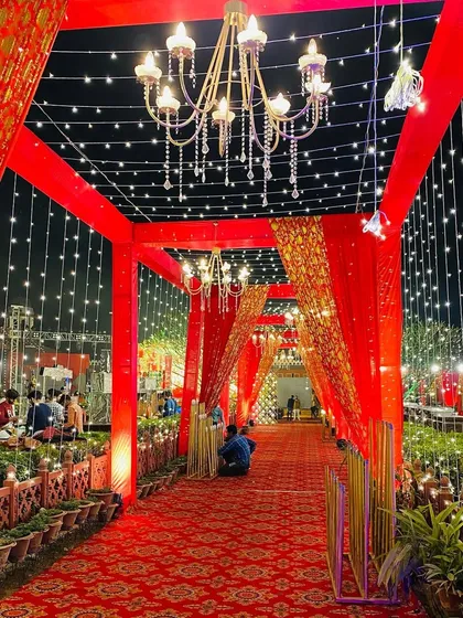 A grand red walkway for an outdoor wedding, framed by structures draped in red and gold fabric and lit by elegant chandeliers.