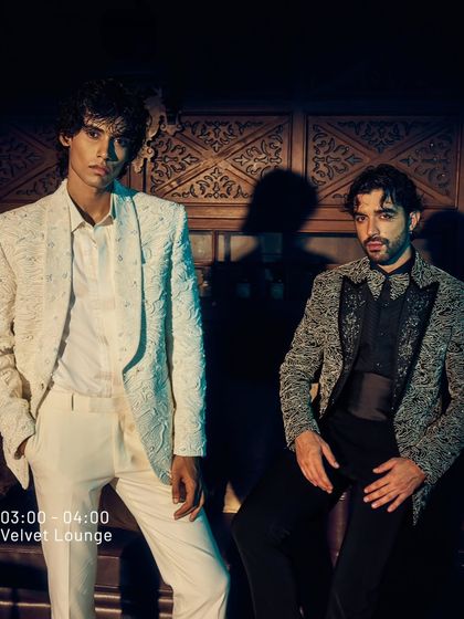 Two men, two statements. One in a pristine white embroidered jacket, the other in a black patterned blazer. This is the duality of the Velvet Lounge, where luxury is lived, not just worn.