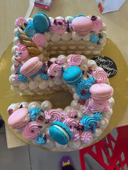 An overhead view of a gorgeous number "5" birthday cake, decorated with swirls of pink and blue frosting and colorful macarons. It's a delicious work of art that makes any birthday feel extra special.
