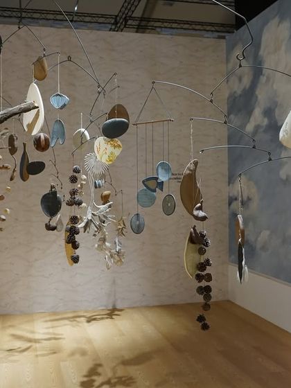 The mobile from the 'Feel More' installation, set against a backdrop of a painted sky. The interplay of the handcrafted mobile, the ethereal fabrics, and the abstract landscape conjures a space where tranquility meets intrigue.