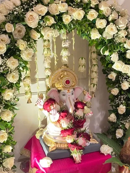 An elegant and pure white rose arch for a serene Ganpati decoration. The hanging bells and fresh green leaves add subtle details to this classic and beautiful design.