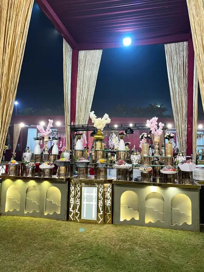 A wide view of an elegant dessert station set up on the lawn. The symmetrical arrangement and soft lighting on the gold and white counters create a sophisticated and inviting display.
