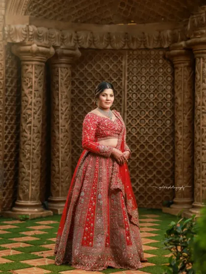 A full-length portrait against intricately carved pillars. The bride's pose is confident and elegant, showcasing the beautiful silhouette of her red lehenga.