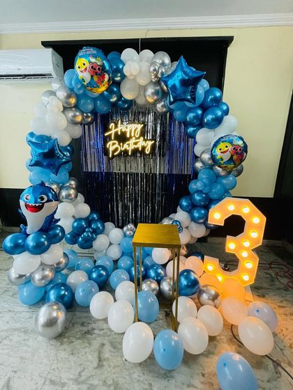A fun Baby Shark theme for a 3rd birthday. The setup includes a blue and white balloon ring, a "Happy Birthday" neon sign, character cutouts, and a light-up number '3'.