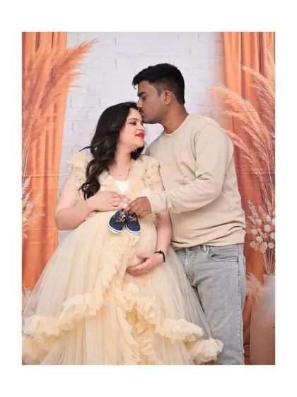 A sweet couple's portrait where the father-to-be is kissing his partner's forehead. They are holding a tiny pair of baby shoes, symbolizing their shared excitement for the new arrival.