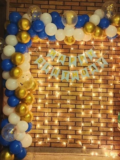 A balloon arch in blue, white, and gold against a brick wall with fairy lights.