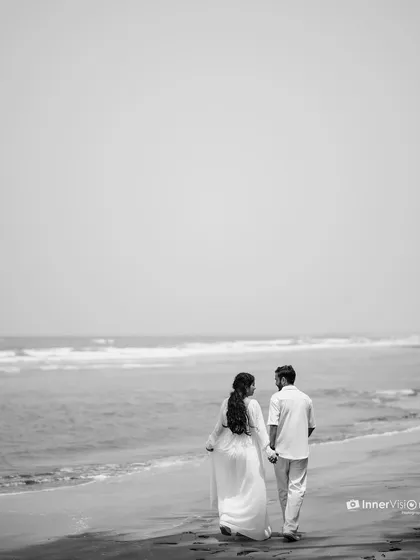 A classic black and white shot of the couple walking away from the camera, hand-in-hand, along the shoreline. The minimalist composition emphasizes their journey together.