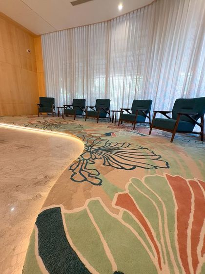 A detail of the GCC Hotel lobby, highlighting the magnificent custom-designed floral carpet. The carpet flows through the space, complemented by modern armchairs and floor-to-ceiling sheer curtains.