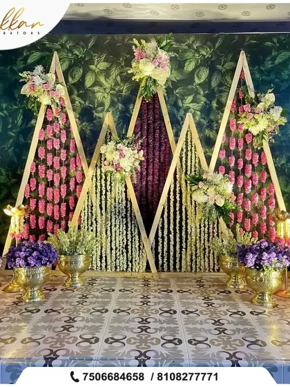 A unique and modern stage backdrop I designed. It features triangular wooden frames filled with hanging strings of pink, purple, and white flowers, creating a geometric and artistic floral wall.