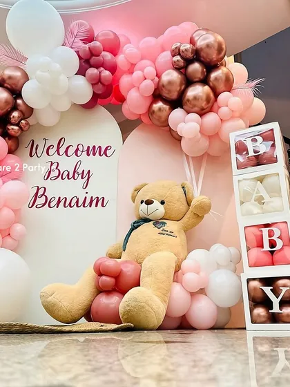 A beautiful and modern welcome home decoration for a baby girl. This setup uses a stylish arch backdrop, a lush balloon arrangement in shades of pink, rose gold, and white, along with "BABY" blocks and a large, cuddly teddy bear.