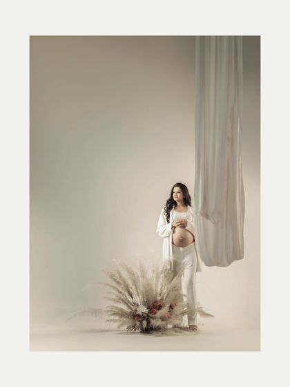A full-length portrait in a minimalist setting. The mother-to-be stands beside a large pampas grass arrangement, her simple white outfit and the neutral tones creating a calm and modern aesthetic.
