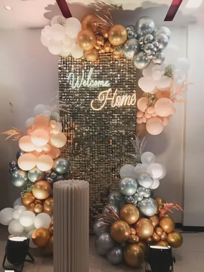 A glamorous welcome home setup featuring a gold sequin wall and a 'Welcome Home' neon sign. The balloon clusters in peach, silver, and gold add a touch of elegance and celebration.