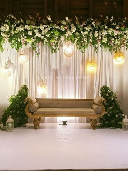 A simple and elegant stage decoration for a Christian wedding. The setup features a white curtain backdrop with an overhead floral arrangement and hanging lanterns.