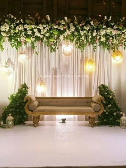 A simple and elegant stage decoration for a Christian wedding. The setup features a white curtain backdrop with an overhead floral arrangement and hanging lanterns.