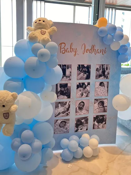 A personalized photo board showcased precious moments, framed by a soft blue balloon arch and topped with a cute teddy bear, adding a deeply personal touch to the baby welcome decor.