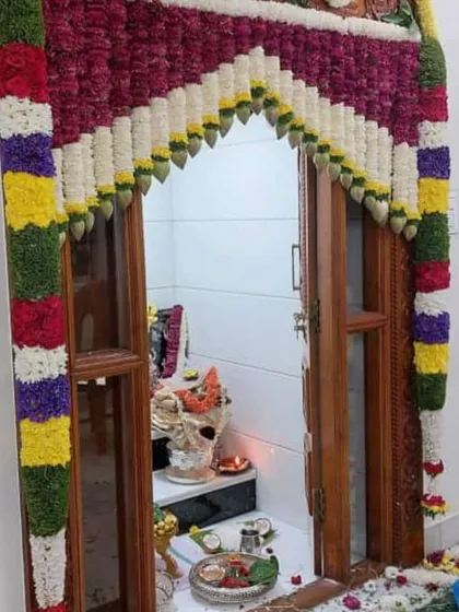 A pooja room entrance decorated with colorful flower garlands. The archway is made with sections of red, white, yellow, green, and purple flowers.