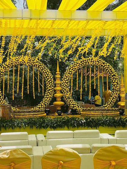 A classic Haldi stage setup with a yellow canopy and large floral rings made of marigolds. The white sofas provide comfortable and stylish seating for the guests.