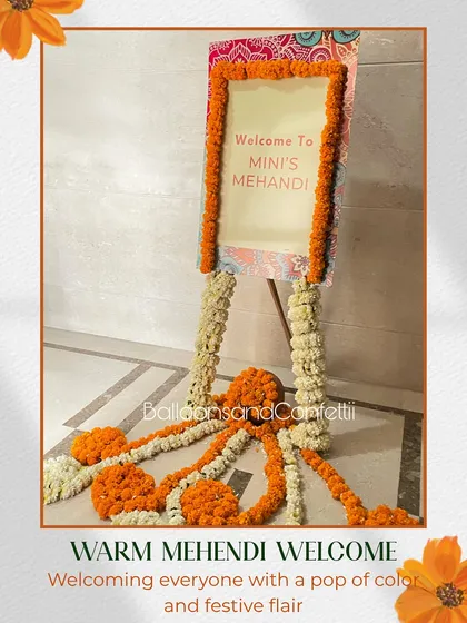 A warm welcome for a Mehendi ceremony. This personalized welcome sign is framed with bright orange marigolds and set on an easel wrapped in white floral garlands, setting a festive tone right from the entrance.