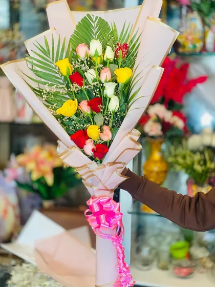 A beautiful hand-tied bouquet of mixed roses in red, yellow, white, and pink. The arrangement is accented with palm leaves and wrapped in pink paper for a classic look.
