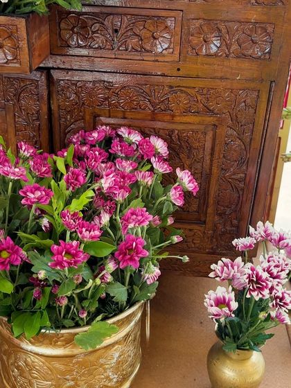 A close-up of pink and white-tipped flowers in a brass pot, set against the warm tones of carved wood.