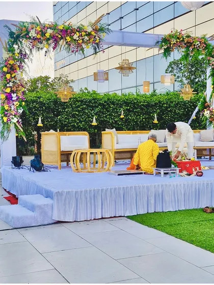 A modern and minimalist vidhi mandap for an outdoor daytime ceremony. The setup features a beautiful floral arch, comfortable cane furniture for the rituals, and elegant hanging lamps.