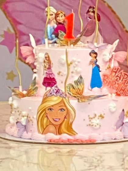 A close-up of a beautiful two-tier princess-themed birthday cake. It features characters like Barbie, Elsa, and other princesses, perfectly matching a fairy-tale party theme.