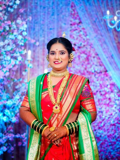 A poised bridal portrait. The bride stands confidently, her hands clasped, showcasing her full wedding attire and traditional gold jewellery against the colorful backdrop.