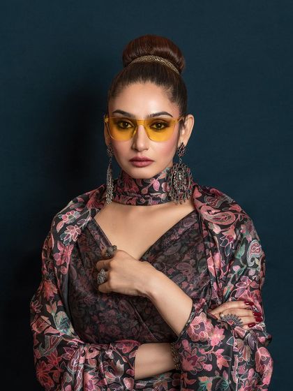 A bold studio portrait of Ragini Dwivedi for a magazine feature. The styling with yellow-tinted glasses and heavy jewelry creates a unique, high-fashion statement.