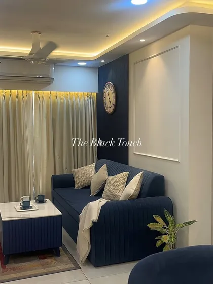 A cozy living room featuring a plush blue sofa against a dark accent wall. The warm, indirect lighting from the false ceiling and the simple decor create a comfortable and relaxing atmosphere, perfect for unwinding after a long day.