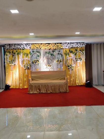 A full view of a stage setup in a banquet hall, ready for a Haldi or Sangeet function. The backdrop features yellow draping and intricate floral work, with a wide red carpet leading up to the main seating area.