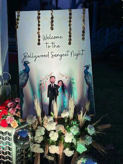 The custom welcome sign for a 'Bollywood Sangeet Night', featuring a caricature of the couple and decorated with pampas grass and disco balls.