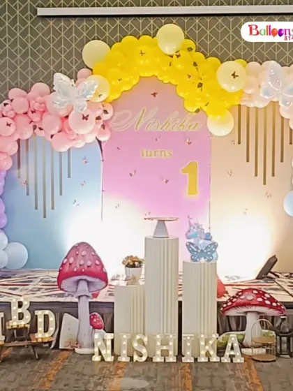 An elegant and whimsical butterfly garden theme for a first birthday. This setup features a beautiful pastel balloon arch, large illuminated butterfly props, and a personalized backdrop for Nishika.