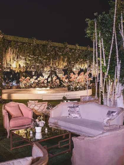 A cozy lounge seating area at a reception, set against a backdrop of a grand stage with oversized paper flowers. The mix of pink and cream furniture creates a soft and romantic corner for guests.