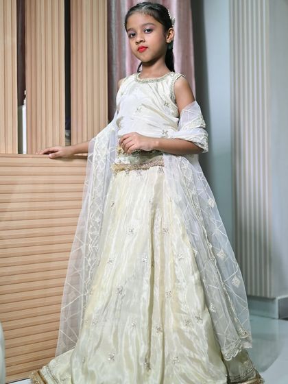 A young model posing indoors in the ivory kids' lehenga. This outfit is available for rent and can be customized to your needs.