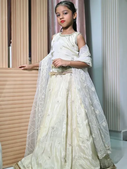 A young model posing indoors in the ivory kids' lehenga. This outfit is available for rent and can be customized to your needs.
