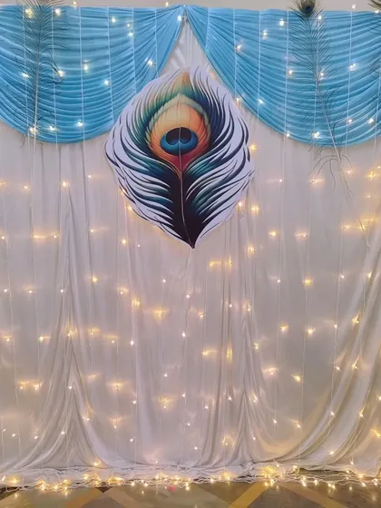 A simple yet elegant backdrop with a large peacock feather motif, white and blue drapes, and fairy lights.