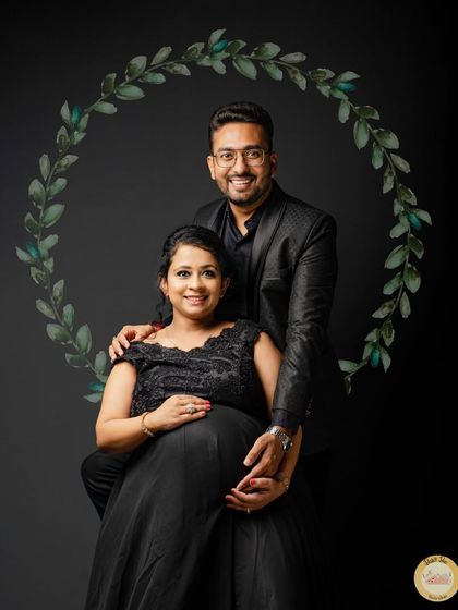 A formal maternity portrait against a solid black background. The couple is dressed in elegant black attire, and a simple digital wreath was added to frame them, creating a classic and sophisticated look.