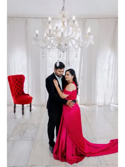 An elegant pre-wedding portrait in a grand, all-white room with a crystal chandelier. The red chair adds a pop of color to this sophisticated studio shoot.