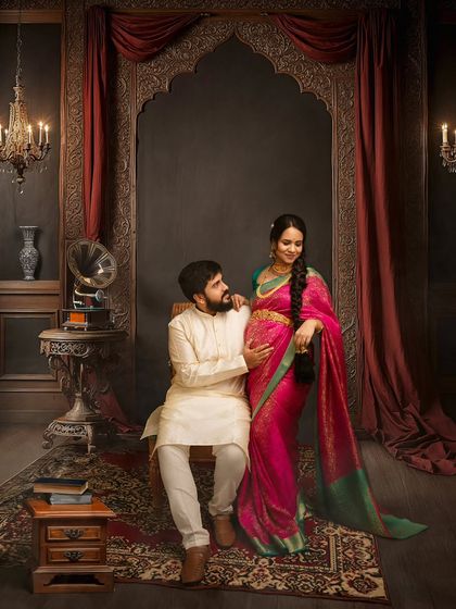 A classic pose showing the couple's bond. The husband-to-be gently holds the baby bump, set against a backdrop of vintage furniture and an ornate archway, creating a truly royal portrait.