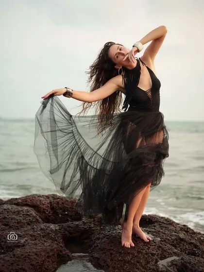 A dynamic pose with the model holding out the sheer black skirt. The movement of the fabric against the backdrop of the sea creates a beautiful and artistic fashion shot.