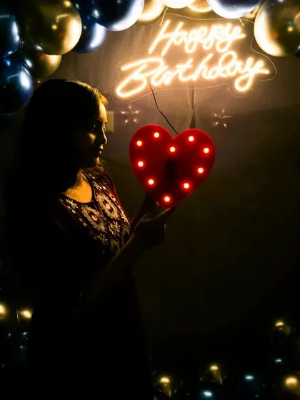 A beautiful silhouette shot of a guest holding a light-up heart prop. The 'Happy Birthday' neon sign and balloon arch create a perfect, softly lit background.