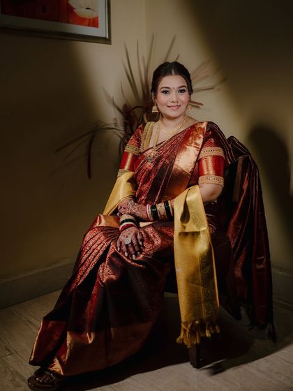 A full-length seated portrait of the bride, looking confidently at the camera. Her rich maroon and gold saree is the star of the shot.