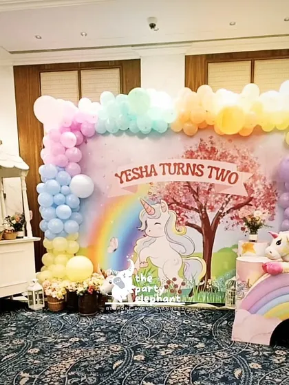This magical unicorn-themed backdrop was designed for a second birthday party. It features a custom illustration of a unicorn under a cherry blossom tree, framed by a beautiful pastel rainbow balloon garland and whimsical props.