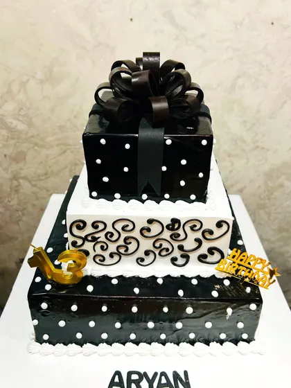 A striking three-tier square cake in a black and white theme. The design includes polka dots, elegant swirls, and a large chocolate bow on top, perfect for a chic 13th birthday celebration.